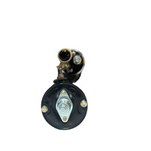 A black automotive part, likely a starter motor or solenoid, positioned on a white surface with a blurred warehouse background.