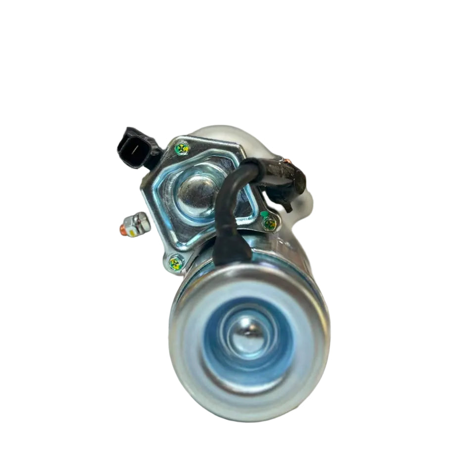 A metal automotive starter motor with a black plastic connector, photographed on a white surface with a blurred warehouse background.
