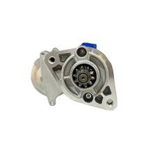 An automotive metal part, likely an alternator or starter motor, displayed on a white counter in a workshop with blurred shelves and a man in the background.